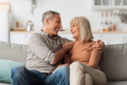 Cheerful Mature Husband Embracing Happy Wife Smiling To Her Sitting On Sofa At Home. Retirement And Family Lifestyle. Romantic Relationship In Senior Age Concept