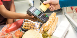 Male consumer paying for food products by credit card while holding it over payment terminal in hand of saleswoman by counter